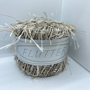 May include: A skein of yarn with a fluffy texture in shades of beige and brown. The yarn is labeled "FLUTTER" on a white label with a silver border.