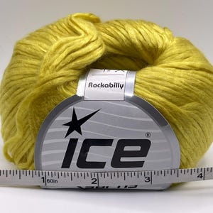 May include: A ball of bright yellow yarn with a paper label reading Ice and Rockabilly. A white measuring tape is positioned in front of the yarn, showing the label width is approximately 3.5 inches across the center of the product.