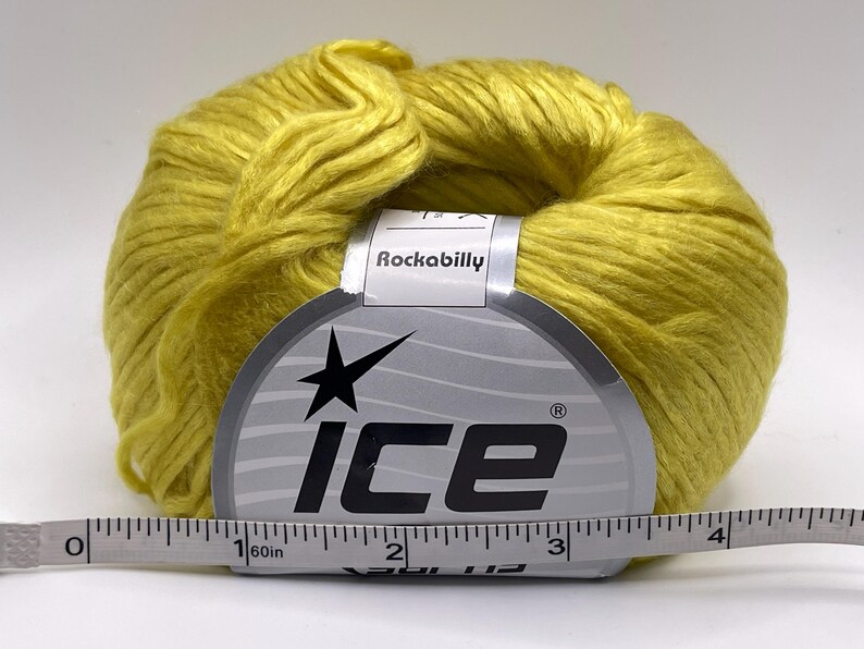 May include: A ball of bright yellow yarn with a paper label reading Ice and Rockabilly. A white measuring tape is positioned in front of the yarn, showing the label width is approximately 3.5 inches across the center of the product.