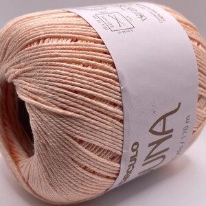 May include: A ball of peach-colored yarn with a white label wrapped around it. The label has the word "UNA" printed on it. The yarn is tightly wound and appears soft. The label also includes measurements and the word "Circulo".