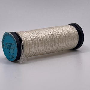 May include: A spool of white metallic thread with a light blue circular label on the end. The label reads Kreinik Metallics, Tapestry (#12) Braids, 100, and 10M. The thread is wound tightly around a black plastic core with black end caps.
