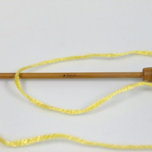 May include: A single light brown wooden knitting needle with a rounded end and the text 4.0mm printed on the shaft. A strand of bright yellow yarn is looped around the needle, extending across the frame in a loose, curved pattern.