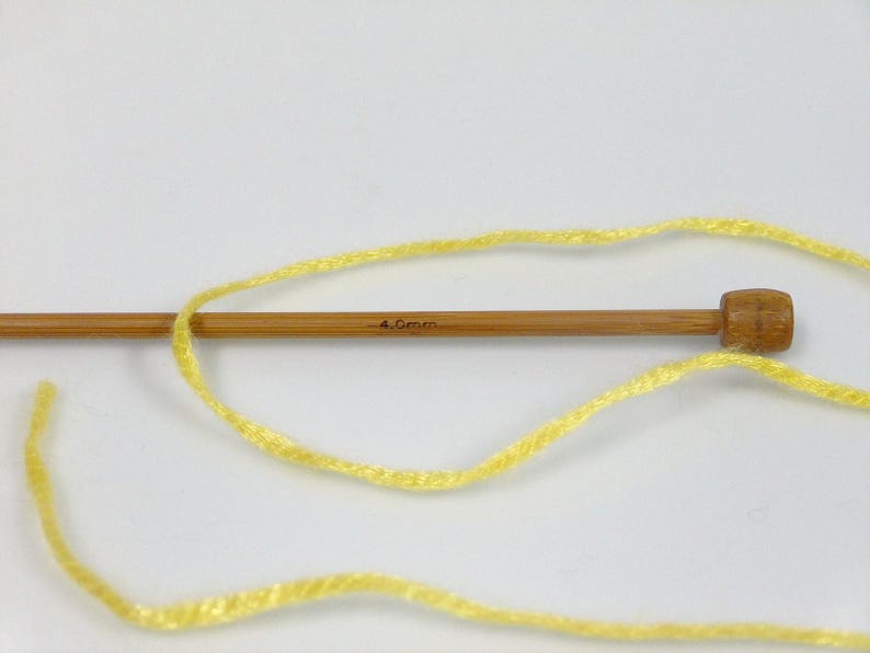 May include: A single light brown wooden knitting needle with a rounded end and the text 4.0mm printed on the shaft. A strand of bright yellow yarn is looped around the needle, extending across the frame in a loose, curved pattern.