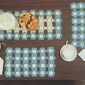 May include: A table setting featuring crocheted placemats and coasters in a blue and white floral pattern. A white teapot, teacups, and a basket of cookies are arranged on the table, along with silverware and a folded napkin.