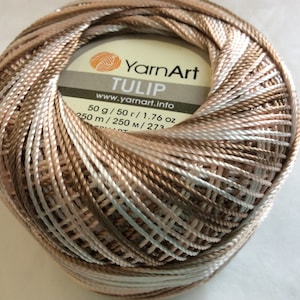 May include: A ball of YarnArt Tulip yarn featuring a variegated colour palette of beige, brown, and white. The centre label displays the brand name YarnArt, the product name TULIP, the website www.yarnart.info, and the weight of 50 g / 1.76 oz.