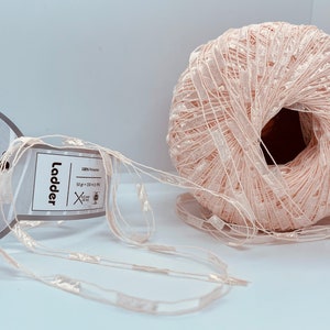 May include: A skein of pale pink yarn with a shimmery finish. The yarn is labeled "Ladder" and is 100% polyester. The skein is 50 grams and 220 meters long.