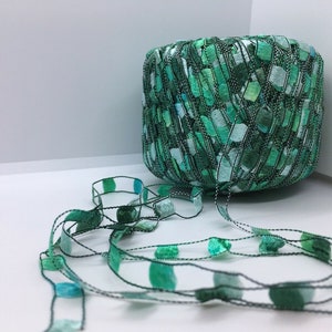 May include: A spool of green and teal ribbon with a textured, woven appearance. The ribbon is a mix of light and dark shades of green, with a black thread outlining the edges. A length of ribbon extends from the spool, featuring teal and green circular accents.