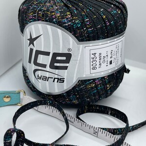 May include: A black ball of yarn with multicoloured glitter threads. The label reads "Ice Yarns", "Namaste Glitz", "80354", "8mm/US 11", "Bulky", "50gr 57m", "84% Polyester 18% Metallic Lurex Black".
