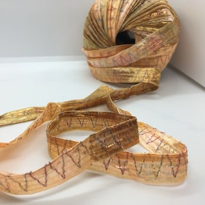 May include: A spool of gold ribbon with a metallic sheen, partially unraveled. The ribbon has a stitched zigzag pattern in dark red and blue thread. The ribbon is approximately 1 inch wide.