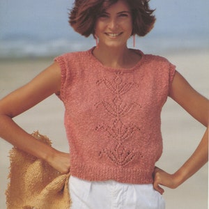 May include: A sleeveless, coral-coloured knitted top with a decorative leaf pattern on the front. The top is paired with white shorts. A straw hat is held in the left hand. The background is a beach scene.