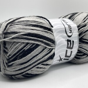 May include: A close-up of two skeins of yarn. The yarn is a blend of black, gray, and white, creating a striped pattern. The label reads "ICE Yarns". This yarn is suitable for knitting and crocheting projects.