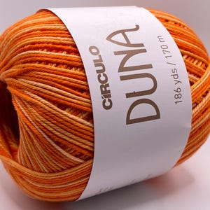 May include: A ball of orange and cream-colored yarn with a white label wrapped around it. The label reads "CIRCULO DUNA" and "186 yds / 170 m". The yarn is tightly wound, showing a gradient of colors.