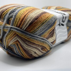 Op de afbeelding: Een bol garen met een mix van kleuren: bruin, beige, geel, grijs en blauw. Het garen is strak opgerold en heeft een etiket aan de zijkant. Geschikt voor brei- of haakprojecten, het garen wordt in gram gemeten.