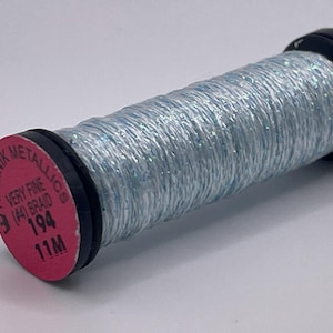 May include: A spool of light blue metallic thread with a shimmering finish. The spool features a pink circular end cap with black text that reads Kreinik Metallics, Very Fine (#4) Braid, 194, and 11M, indicating the product type and length.
