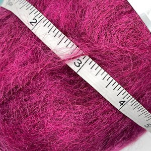 May include: A ball of vibrant magenta yarn is measured with a white measuring tape. The tape shows a measurement of approximately 3 inches. The yarn has a soft, fuzzy texture, ideal for knitting or crocheting projects.