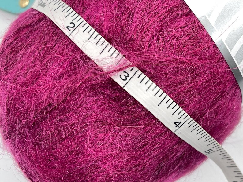 May include: A ball of vibrant magenta yarn is measured with a white measuring tape. The tape shows a measurement of approximately 3 inches. The yarn has a soft, fuzzy texture, ideal for knitting or crocheting projects.