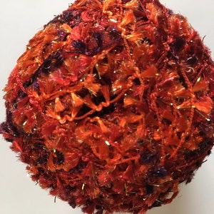 May include: A ball of textured yarn in shades of orange, red, and purple, with gold metallic threads. The yarn has a fuzzy, tassel-like appearance, suitable for knitting or crochet projects. The ball is round and sits against a white background.