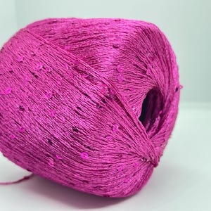 May include: A ball of bright fuchsia yarn with embedded sequins. The yarn is tightly wound, creating a textured surface. The sequins are scattered across the surface, adding a touch of sparkle. Suitable for knitting and crochet projects.
