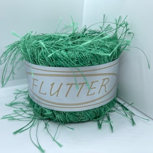 May include: A ball of mint green decorative raffia with the word "FLUTTER" printed on a white label with gold trim. The raffia has a textured, shredded appearance, with loose strands extending outwards.