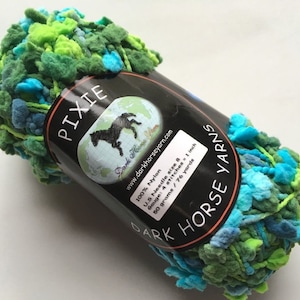以下が含まれることがあります： 緑、青、ターコイズの糸の玉。ラベルには「PIXIE Dark Horse Yarns」と記載されており、糸の素材、針のサイズ、糸の長さなどの情報が記載されています。