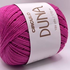 May include: A vibrant magenta yarn ball with a white label wrapped around it. The label reads "CIRCULO DUNA" and "186 yds / 170 m". The yarn is tightly wound, showing the texture of the fibers. Ideal for knitting and crochet projects.