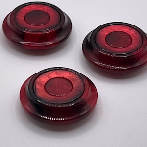 May include: Three red, circular objects with a layered design. Each object has a dark red center and a lighter red outer ring, with a black border. The objects are arranged on a white surface.