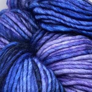 Op de afbeelding: Close-up van een streng garen in tinten blauw en paars. Het garen lijkt dik en getextureerd, met kleurvariaties die diepte cre&euml;ren. De kleuren vari&euml;ren van diep indigo tot lichtere tinten lavendel. Ideaal voor brei- of haakprojecten.