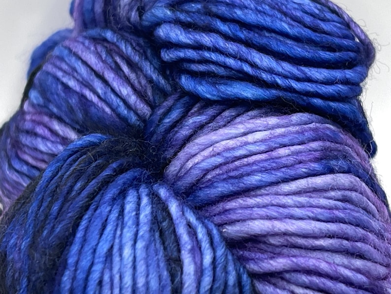 Op de afbeelding: Close-up van een streng garen in tinten blauw en paars. Het garen lijkt dik en getextureerd, met kleurvariaties die diepte cre&euml;ren. De kleuren vari&euml;ren van diep indigo tot lichtere tinten lavendel. Ideaal voor brei- of haakprojecten.