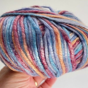 May include: A round ball of variegated yarn featuring a blend of soft blue, purple, peach, and magenta fibers. The yarn has a smooth, slightly lustrous texture with multiple colors twisted together to create a continuous, multi-toned appearance.