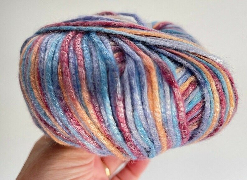 May include: A round ball of variegated yarn featuring a blend of soft blue, purple, peach, and magenta fibers. The yarn has a smooth, slightly lustrous texture with multiple colors twisted together to create a continuous, multi-toned appearance.