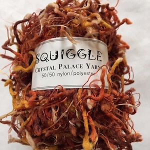 Puede incluir: Ovillo de lana Squiggle Crystal Palace en tonos marrón, naranja y dorado. El hilo es una mezcla de 50/50 nailon y poliéster. La etiqueta dice "Squiggle Crystal Palace Yarn". El hilo tiene un aspecto texturizado y rizado.