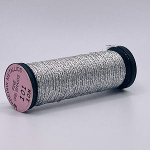 May include: A spool of silver metallic thread with black end caps. The pink label on the end reads KREINIK METALLICS, FINE (#8) BRAIDS, 101, 10M. The thread is tightly wound around the cylindrical spool, creating a textured, shimmering surface.
