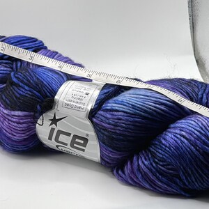 Op de afbeelding: Een streng handgeverfde merinowol in tinten blauw, paars en zwart. Een witte meetlint ligt over het garen en toont metingen in centimeters. Het garenlabel luidt "ice yarns".