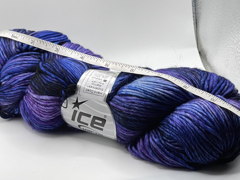 Op de afbeelding: Een streng handgeverfde merinowol in tinten blauw, paars en zwart. Een witte meetlint ligt over het garen en toont metingen in centimeters. Het garenlabel luidt "ice yarns".