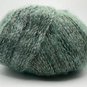 May include: A ball of soft, green yarn with flecks of white, silver, and brown. The yarn has a fluffy texture, ideal for knitting or crocheting projects. The yarn is a muted green color.