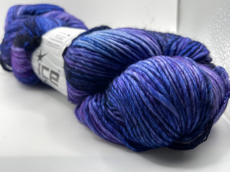 Op de afbeelding: Een bol garen in tinten blauw en paars. Het garen is strak gewikkeld, met zichtbare strengen en een lichte glans. Een wit label met het woord "ice" is zichtbaar.