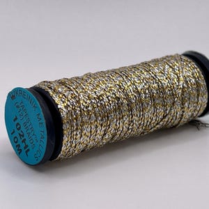 May include: A spool of Kreinik metallic tapestry braid thread featuring a twisted blend of gold and silver metallic fibers. The blue circular label on the end displays the text Kreinik Metallics, Tapestry (#12) Braid, 102HL, and 10M.