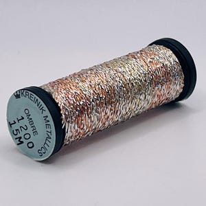 May include: A spool of Kreinik metallic thread featuring a multi-colored ombre blend of silver, copper, and gold tones. The spool ends are black, and the label on the end reads KREINIK METALLICS, OMBRE, 1200, 15M. The thread has a reflective texture.