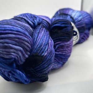 Op de afbeelding: Een close-up van een bol garen in blauwe en paarse tinten. Het garen is dik en getextureerd, met een donkerblauwe sectie in het midden. De kleuren vloeien in elkaar over, waardoor een gemarmerd effect ontstaat. Het garen is waarschijnlijk voor breien of haken.