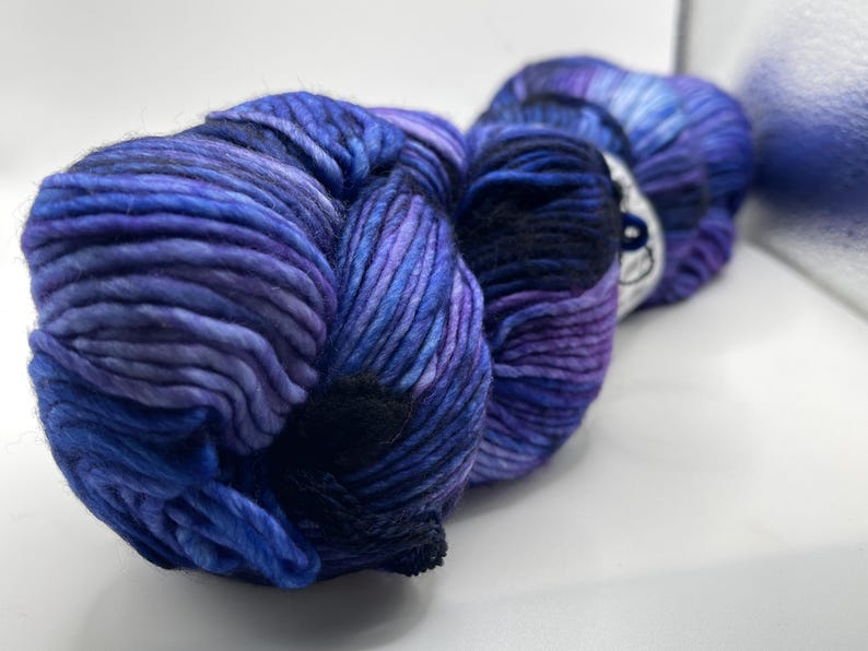 Op de afbeelding: Een close-up van een bol garen in blauwe en paarse tinten. Het garen is dik en getextureerd, met een donkerblauwe sectie in het midden. De kleuren vloeien in elkaar over, waardoor een gemarmerd effect ontstaat. Het garen is waarschijnlijk voor breien of haken.