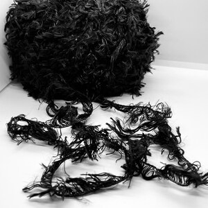 May include: A black ball of yarn with long, shaggy strands. The yarn is unraveling and the strands are spread out on a white surface.