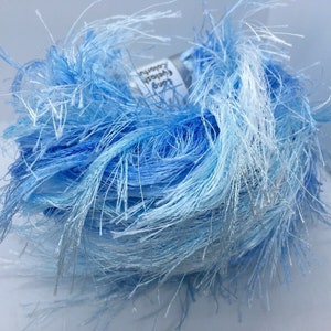 May include: A ball of blue eyelash yarn with varying shades of light and dark blue. The yarn has a fluffy, textured appearance with long, wispy fibers extending outwards. This yarn is suitable for knitting and crochet projects.