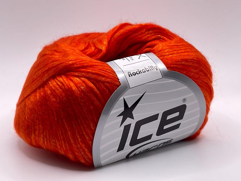 Puede incluir: Ovillo de lana naranja brillante con una etiqueta que dice "Rockabilly" y "ice yarns". La etiqueta tiene un dise&ntilde;o plateado y negro. La lana es de color liso, adecuada para proyectos de punto o ganchillo. La lana es de color naranja brillante.