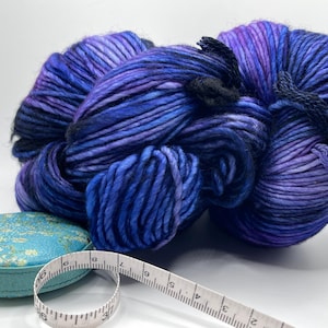 Op de afbeelding: Een grote streng handgeverfde wol in blauwe en paarse tinten, met zwarte banden. Een kleine, turquoise, ronde container en een wit meetlint staan op de voorgrond. Het garen is voor brei- of haakprojecten.