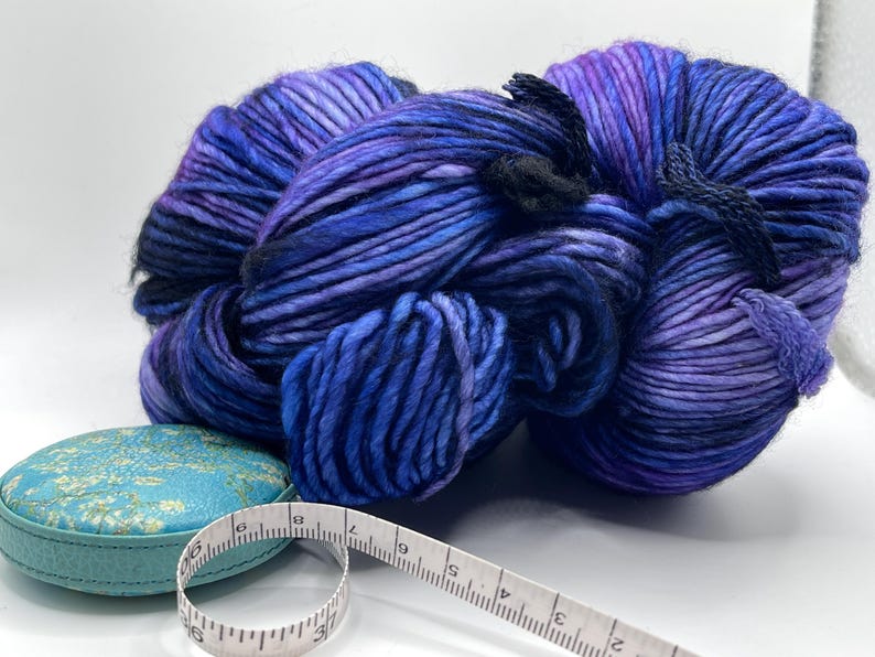 Op de afbeelding: Een grote streng handgeverfde wol in blauwe en paarse tinten, met zwarte banden. Een kleine, turquoise, ronde container en een wit meetlint staan op de voorgrond. Het garen is voor brei- of haakprojecten.
