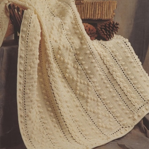 May include: A crocheted blanket with a textured pattern in a cream color. The blanket is folded and draped over a wooden surface.