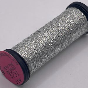 May include: A spool of silver metallic thread with a black core and a pink label on the end. The label reads KREINIK METALLICS, VERY FINE (#4) BRAID, 3231, and 11M. The thread is tightly wound around the cylindrical spool in a braided pattern.