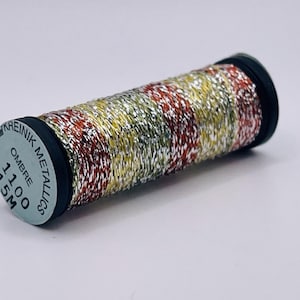 May include: A spool of Kreinik metallic thread featuring an ombre color pattern of silver, gold, red, and green. The label on the end of the black spool reads Kreinik Metallics, Ombre, 1100, and 15M. The thread has a reflective, shiny texture.