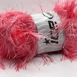 May include: A close-up of a ball of pink yarn with a silver label. The yarn is fluffy and textured, with long, thin strands extending outwards. The label has the letters "JB" and a star.