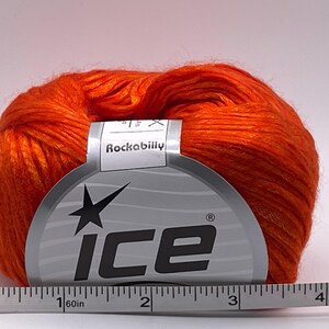 Puede incluir: Una madeja de hilo naranja brillante con una etiqueta "Rockabilly" y la marca "ICE". Una cinta m&eacute;trica debajo muestra una longitud de aproximadamente 10 cm.
